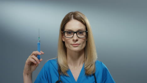 Portrait-of-the-Caucasian-young-blond-beautiful-woman-physician-in-the-glasses-holding-a-syringe-with-a-needle-and-smiling-cherfully.-Close-up.